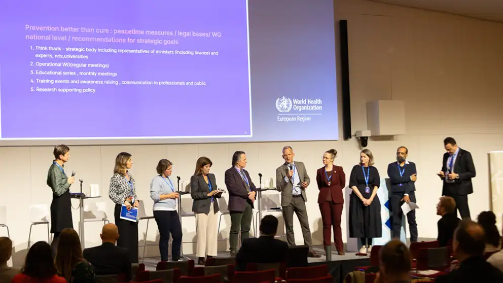 Ten people are presenting on a stage with a Power Point screen in the backgroup. A man is holding a microphone.
