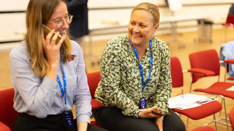 Women smiling. One of them is speaking on a mobile phone, the other one is looking a her.